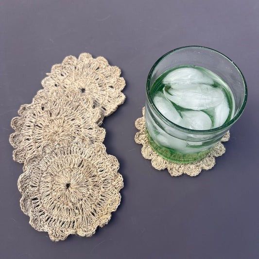 Hand-crocheted nettle coasters in beige color, displayed on a table with a glass of water and ice.