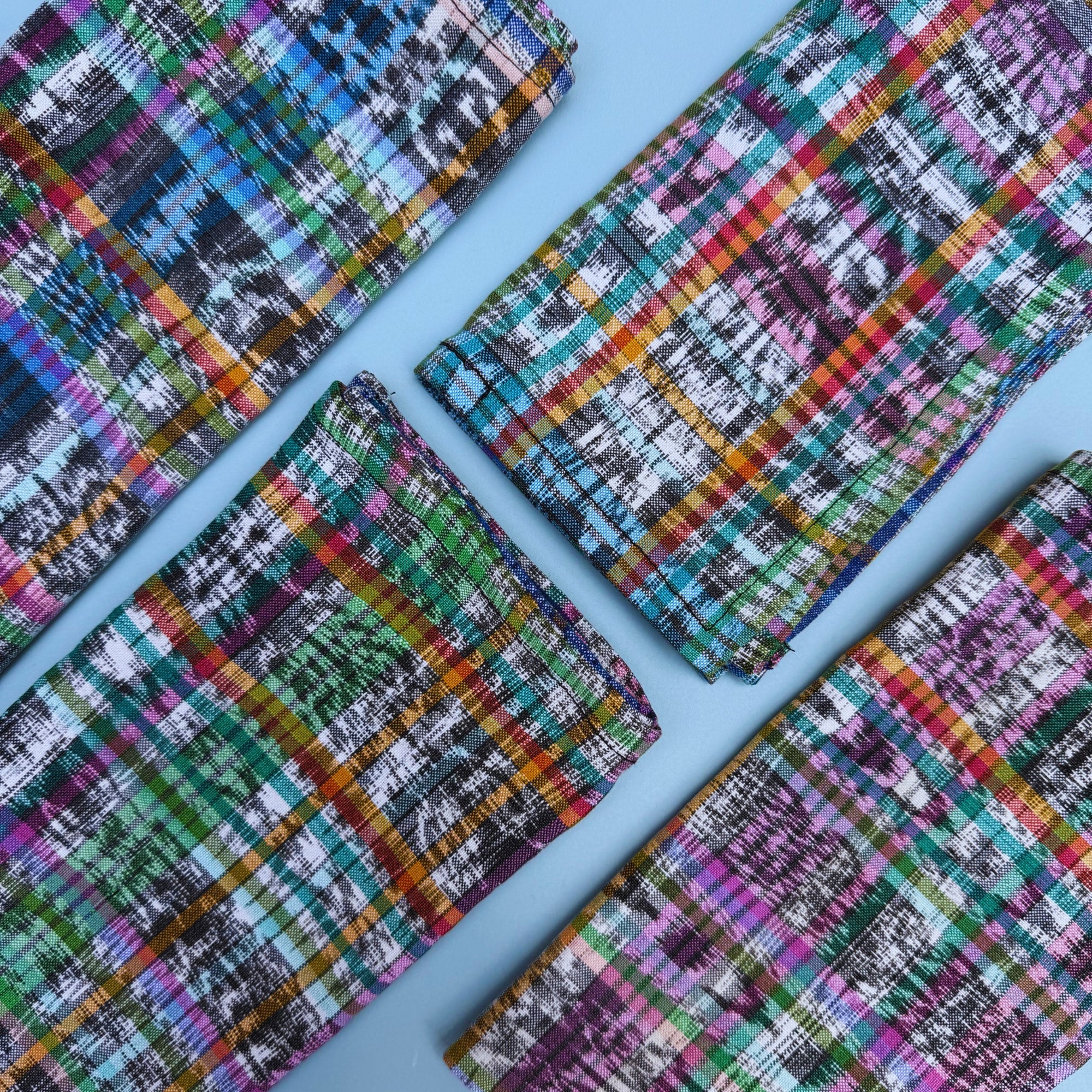 Four folded cloth napkins with a vibrant multicolor plaid ikat pattern, arranged diagonally on a solid light blue background.