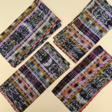 Four folded rectangular cloth napkins with a multicolor plaid and ikat pattern, arranged diagonally on a light beige background.