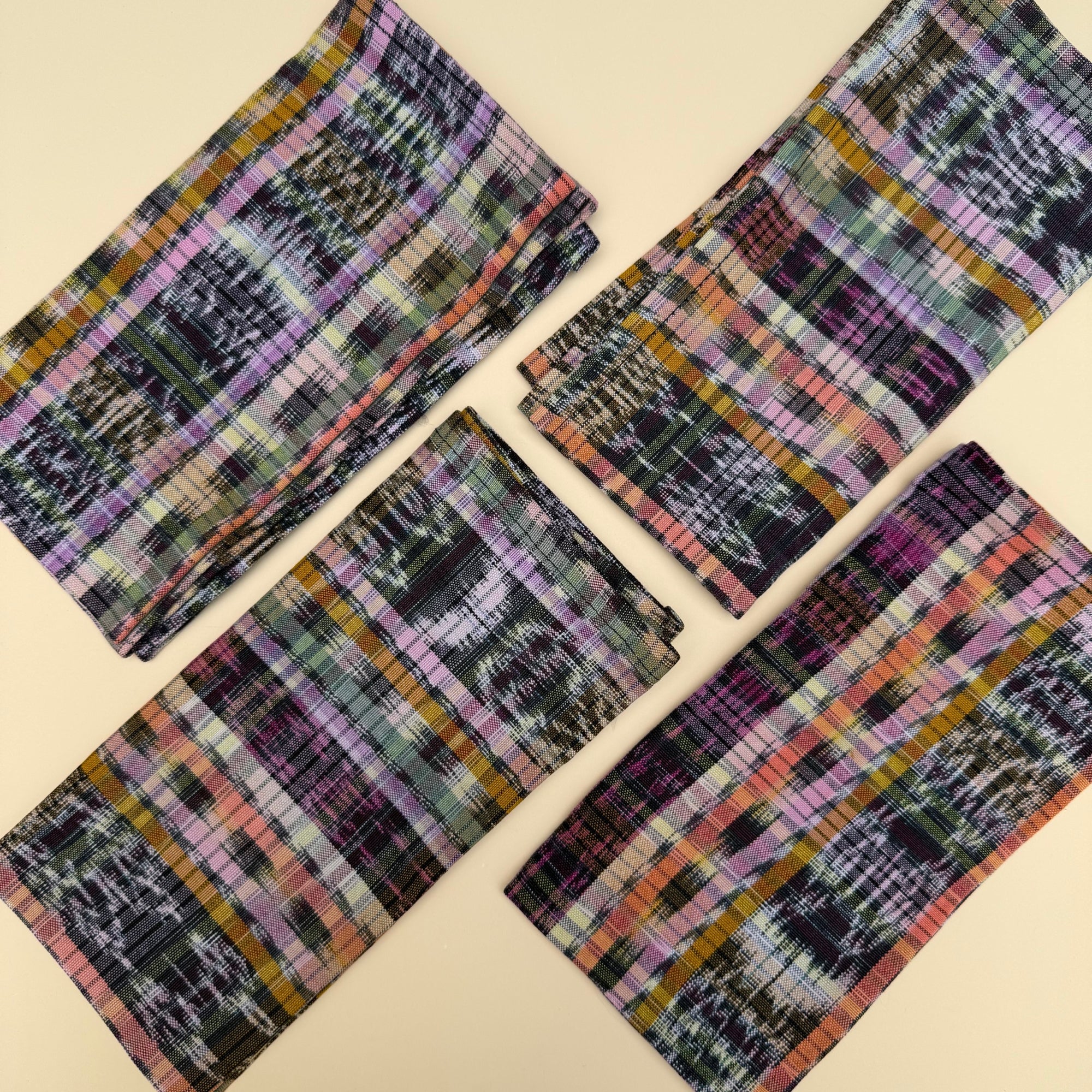 Four folded rectangular cloth napkins with a multicolor plaid and ikat pattern, arranged diagonally on a light beige background.