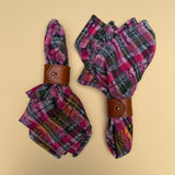 Two folded napkins with a pink, olive green, orange and black plaid pattern, each wrapped in a brown leather napkin ring, displayed on a solid yellow-beige background.