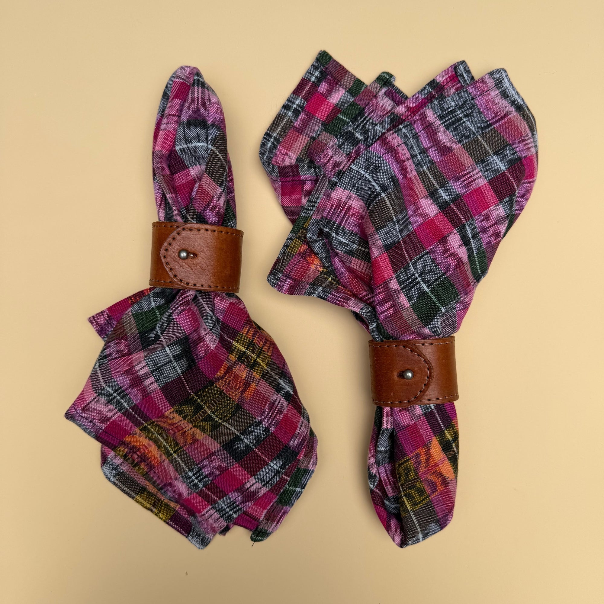 Two folded napkins with a pink, olive green, orange and black plaid pattern, each wrapped in a brown leather napkin ring, displayed on a solid yellow-beige background.