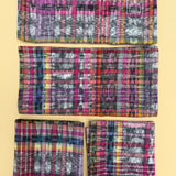 Four folded woven napkins with a colorful plaid and ikat pattern, arranged vertically on a solid pale yellow background.