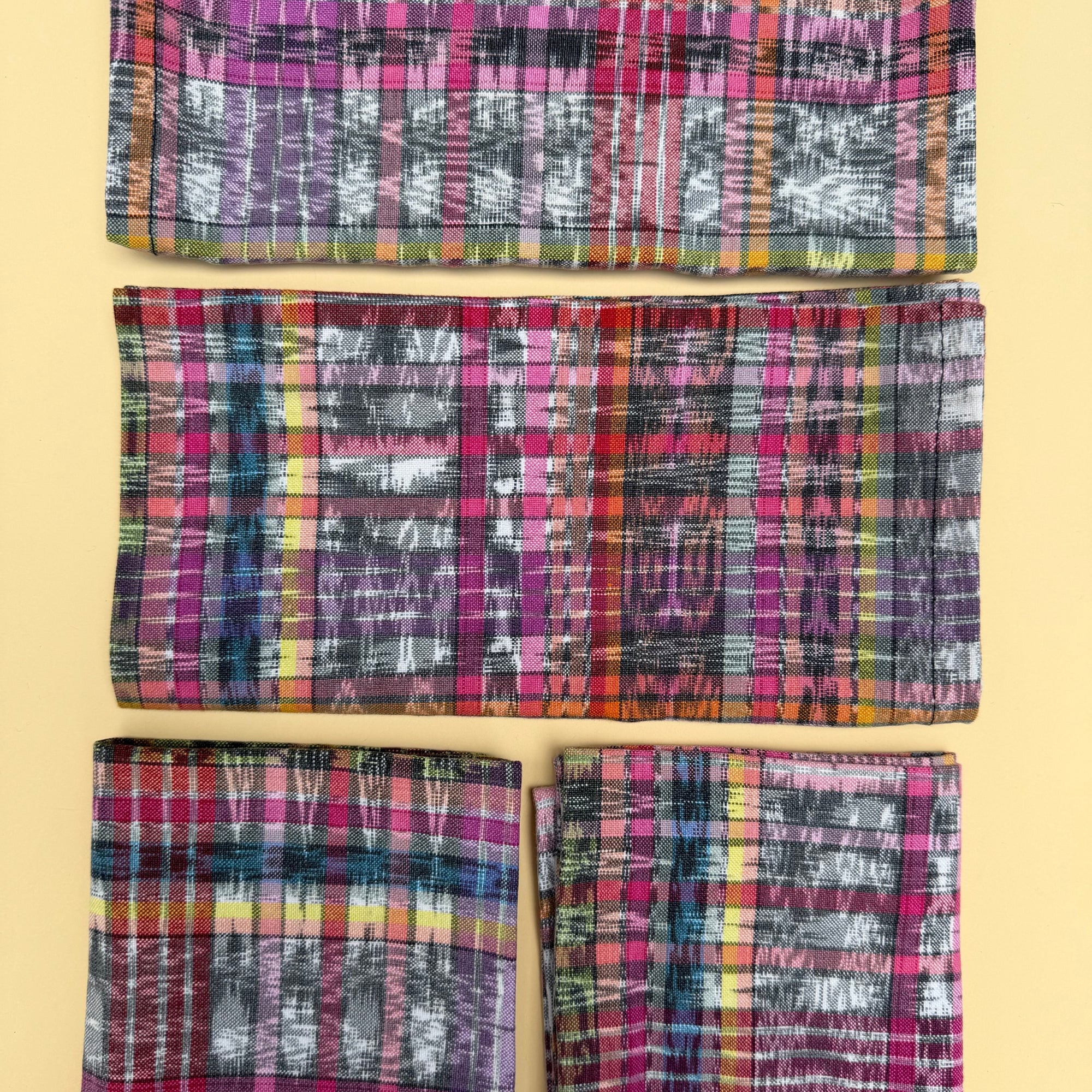 Four folded woven napkins with a colorful plaid and ikat pattern, arranged vertically on a solid pale yellow background.