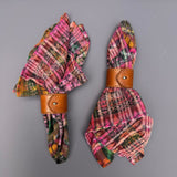 Two folded plaid napkins in pink, orange, green, and yellow tones, each secured with a brown leather ring on a plain gray background.