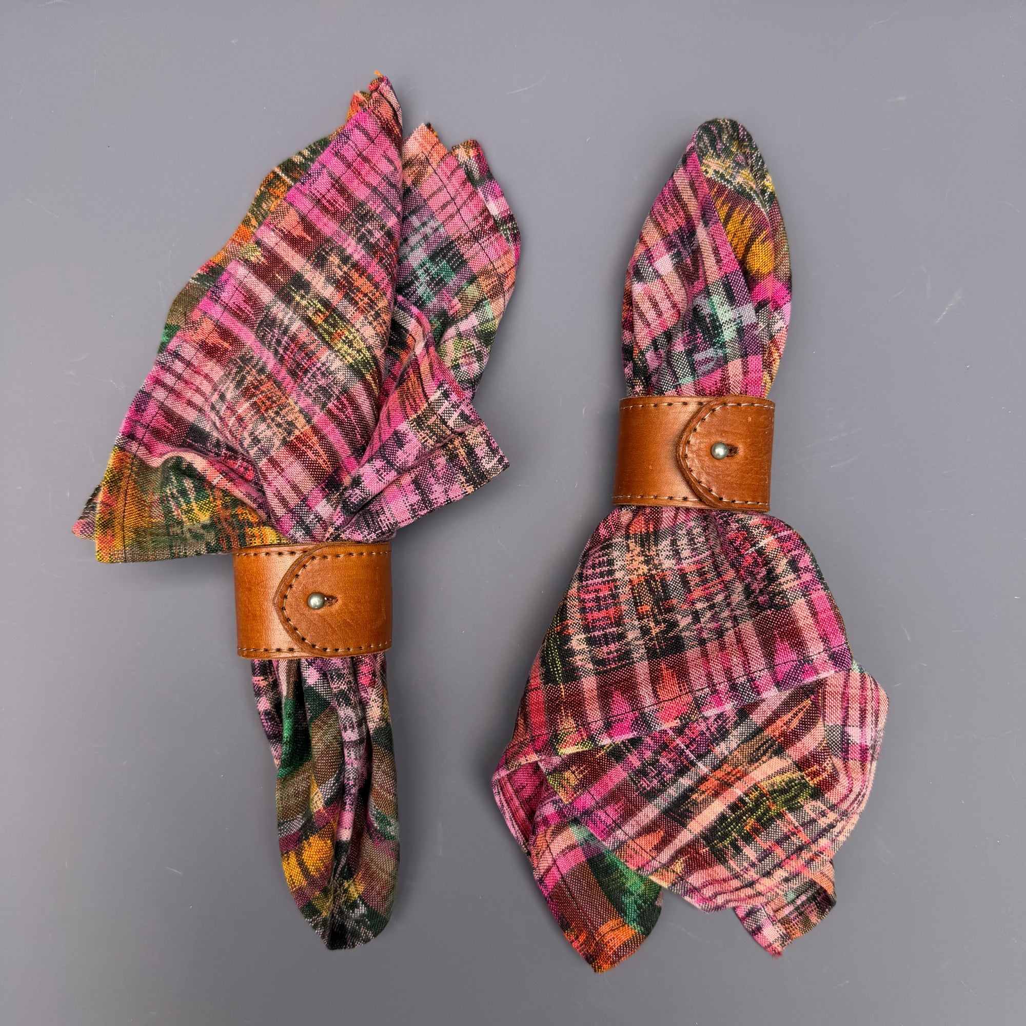 Two folded plaid napkins in pink, orange, green, and yellow tones, each secured with a brown leather ring on a plain gray background.