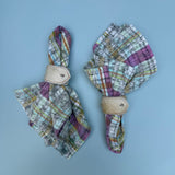 Two fabric napkins folded into cream hair-on-hide napkin rings with metal snap buttons, displayed on a light blue background. The napkins feature a multicolor plaid ikat pattern with purple, green, blue, and white tones.