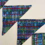 Three triangularly folded napkins with a colorful plaid pattern in blue, green, red, and purple, arranged on a light beige background.