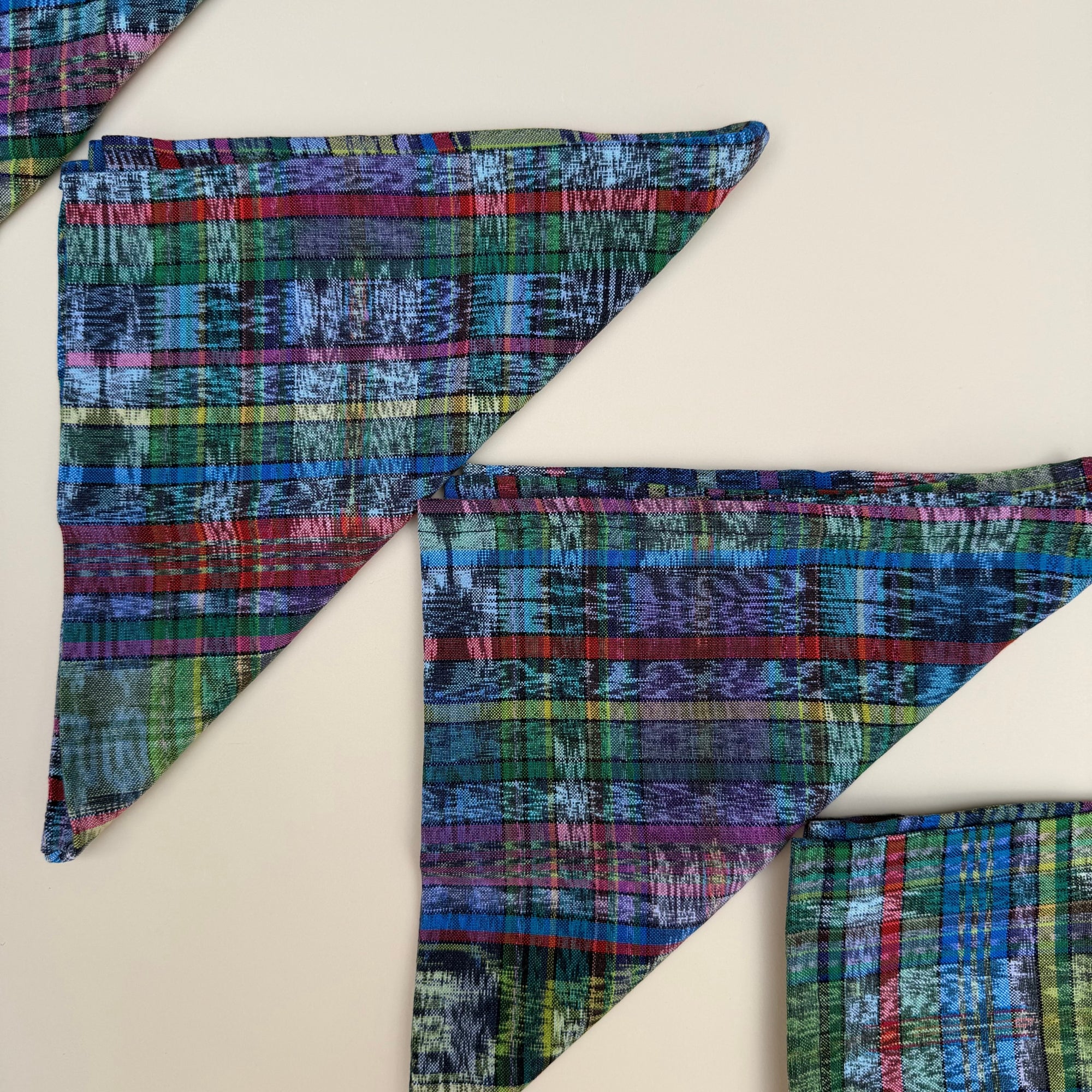 Three triangularly folded napkins with a colorful plaid pattern in blue, green, red, and purple, arranged on a light beige background.