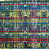 A close-up of a woven textile with a dense multicolor plaid and ikat pattern, featuring stripes of blue, green, red, yellow, purple, and black with white motifs.
