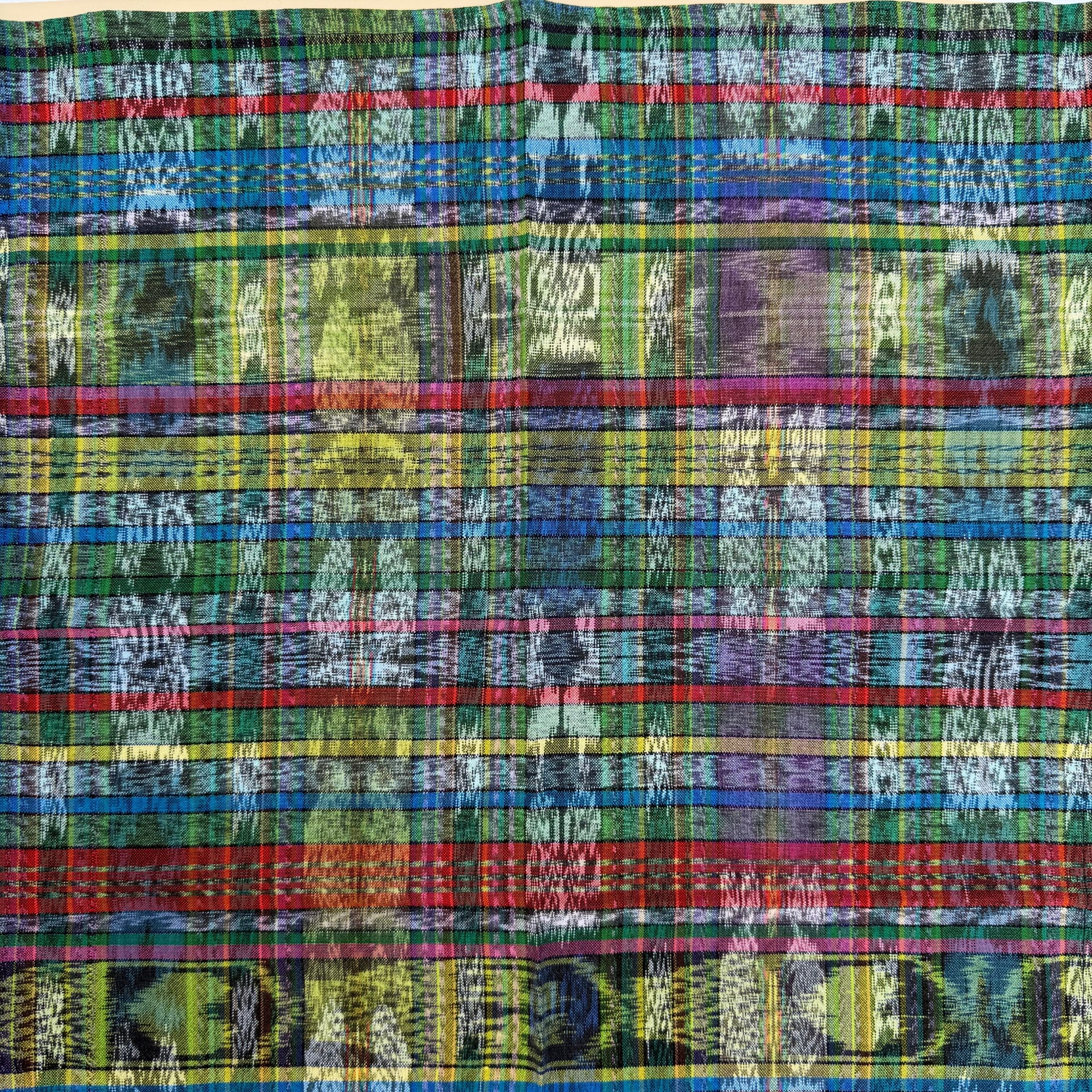 A close-up of a woven textile with a dense multicolor plaid and ikat pattern, featuring stripes of blue, green, red, yellow, purple, and black with white motifs.