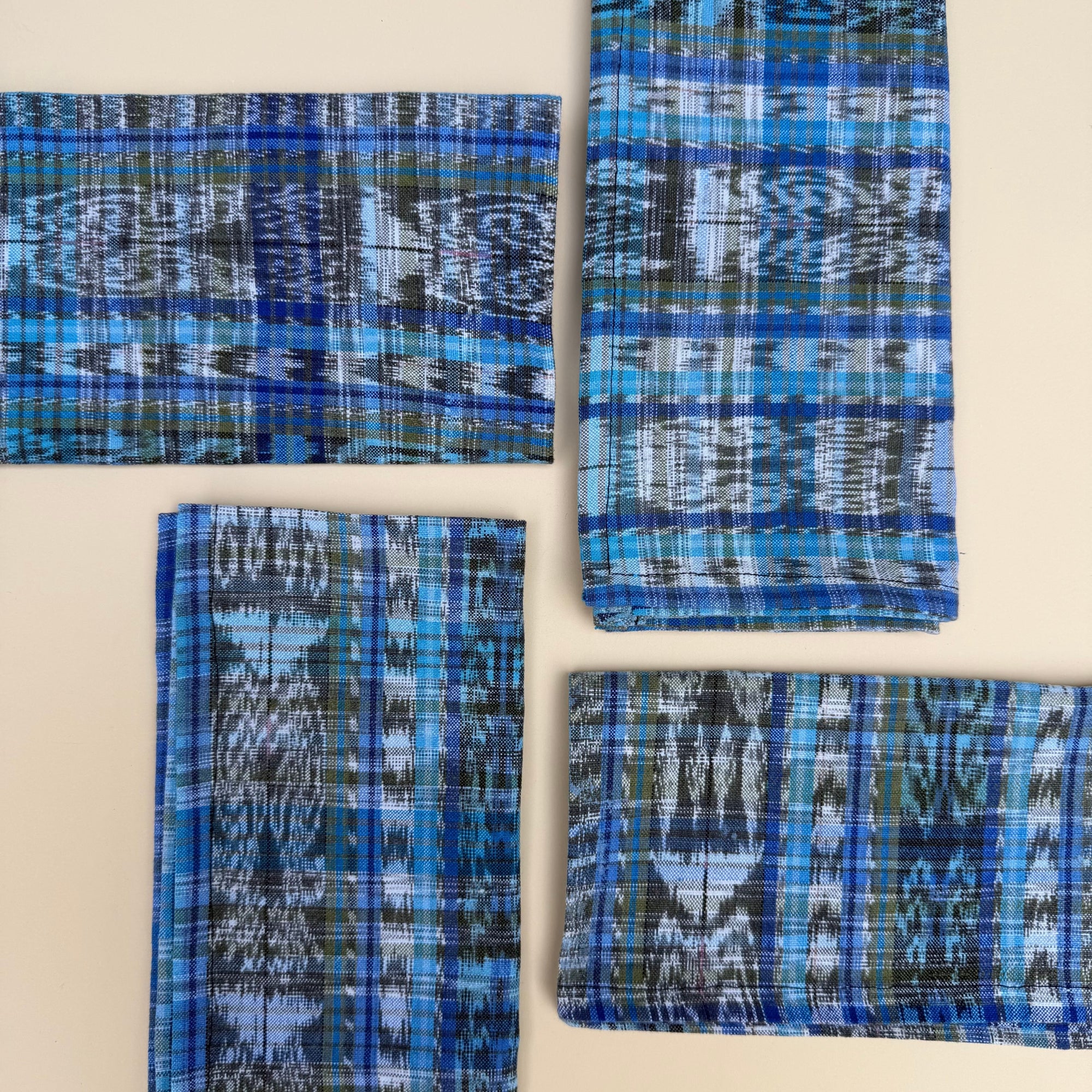 Four folded napkins with blue, navy, green, and white ikat patterns, arranged on a light beige background.