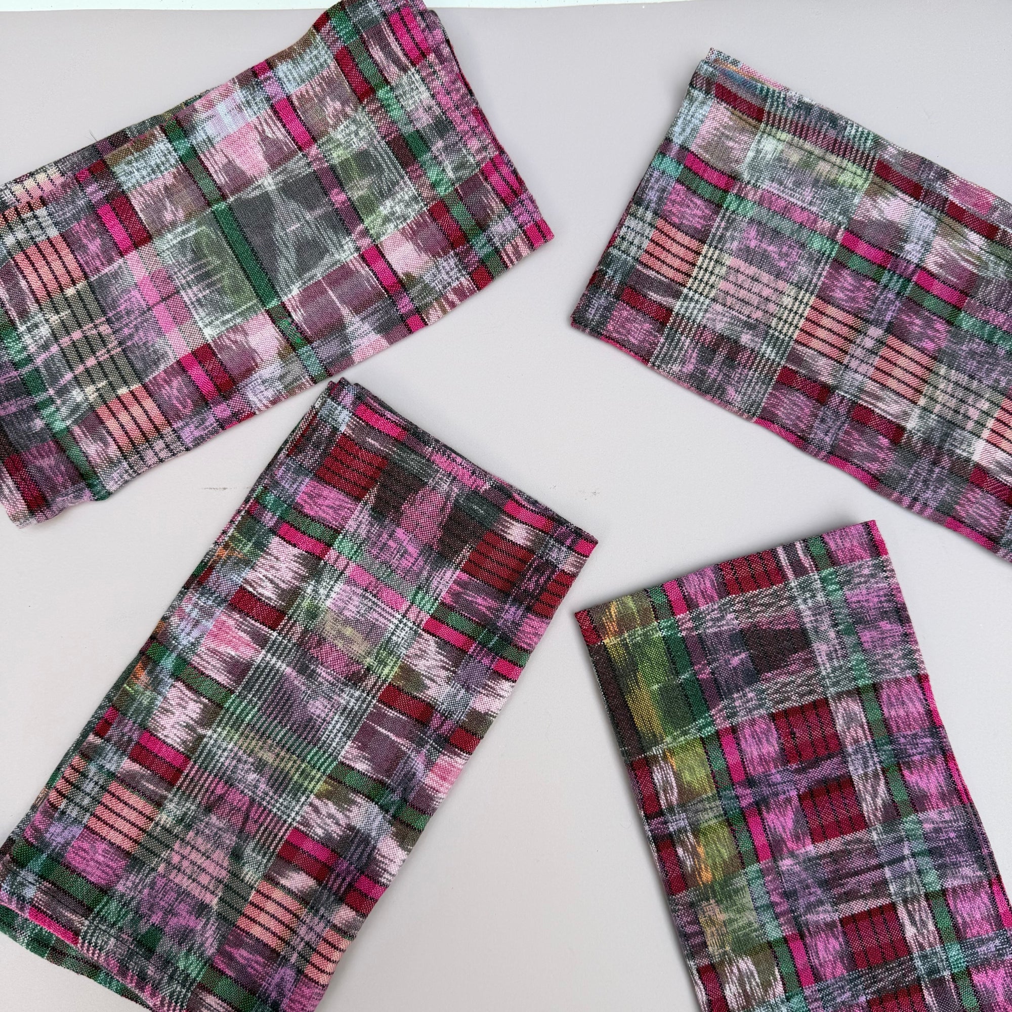 Four rectangular fabric napkins with a colorful plaid ikat pattern in pink, green, red, and white, arranged on a plain white background.