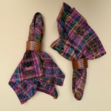 Two folded napkins with a colorful plaid ikat pattern in purple, pink, blue, green, and orange, each secured with a brown leather napkin ring, displayed on a solid beige background.