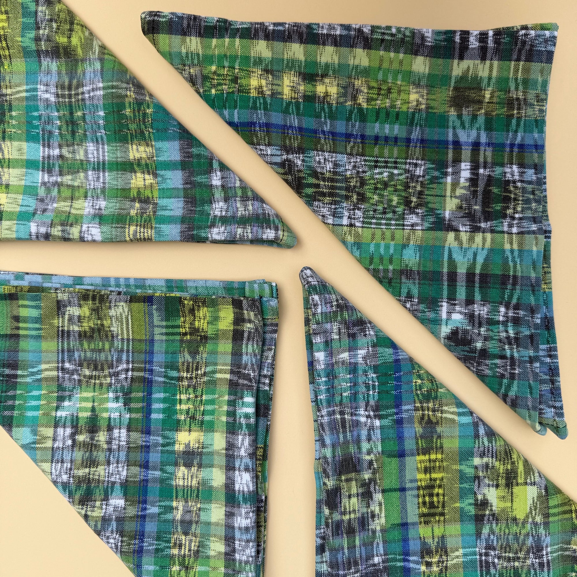 Four cloth napkins, folded in triangles,  with a green, blue, and yellow plaid pattern arranged on a beige background.