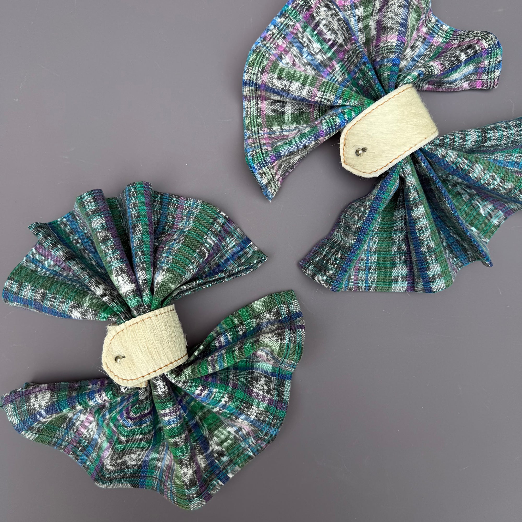 Two fabric napkins with a green, blue, and purple plaid ikat pattern folded accordian style in cream hair-on-hide napkin rings, displayed on a solid gray background.
