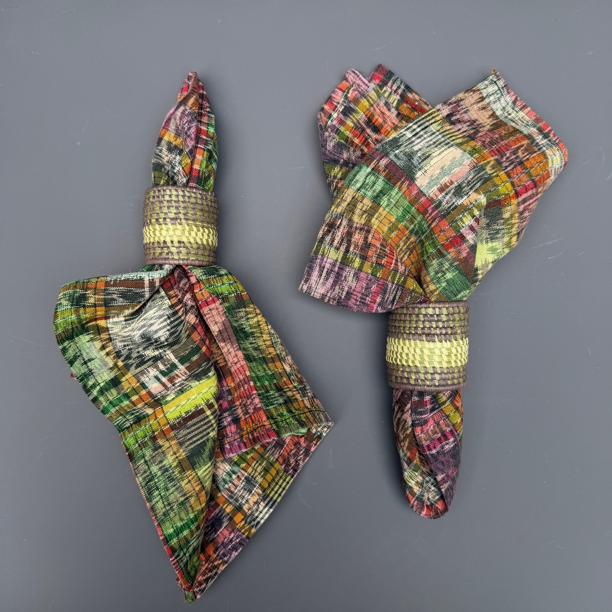 Two folded multicolor plaid ikat napkins with woven beige and yellow bands, arranged on a dark gray background.