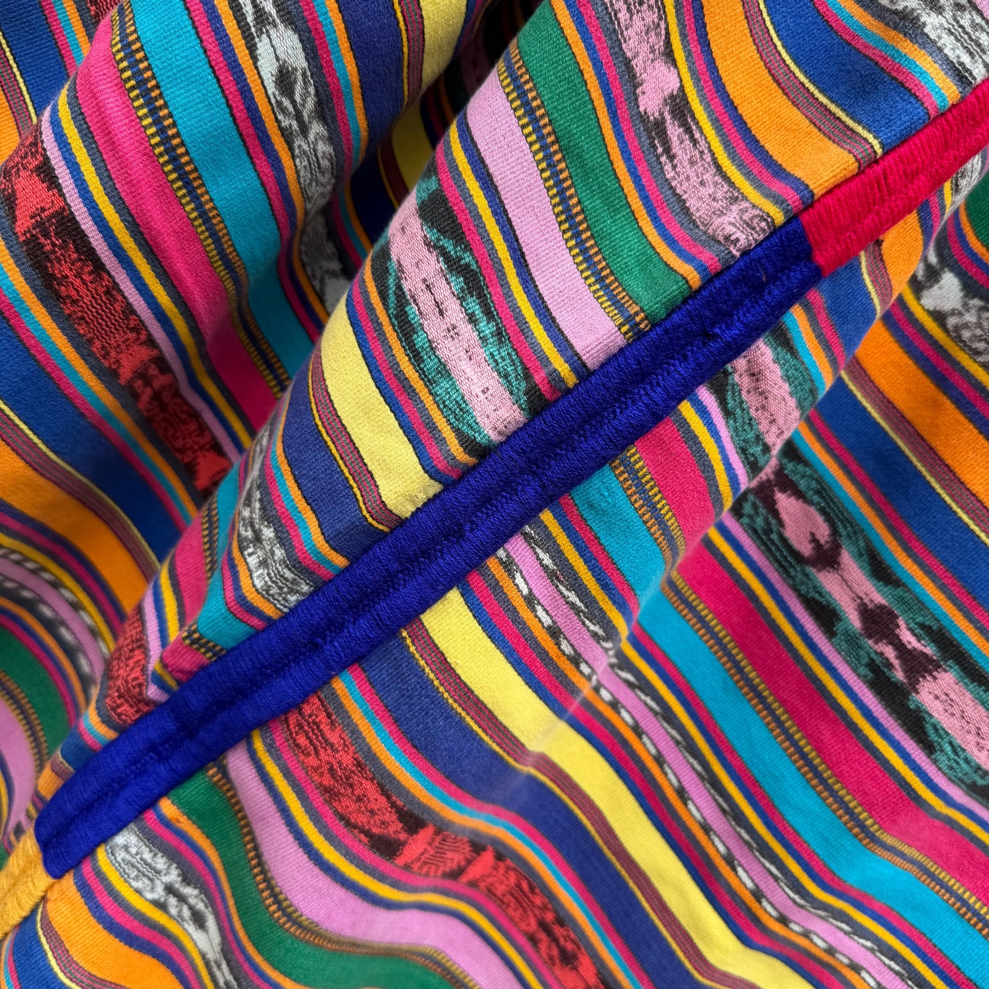 Close-up of a vibrant, multicolored striped fabric with a thick royal blue embroidery running diagonally across the frame.