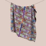 A cotton blanket with a vibrant multicolor geometric pattern, hanging from a thin wire against a solid light beige background.