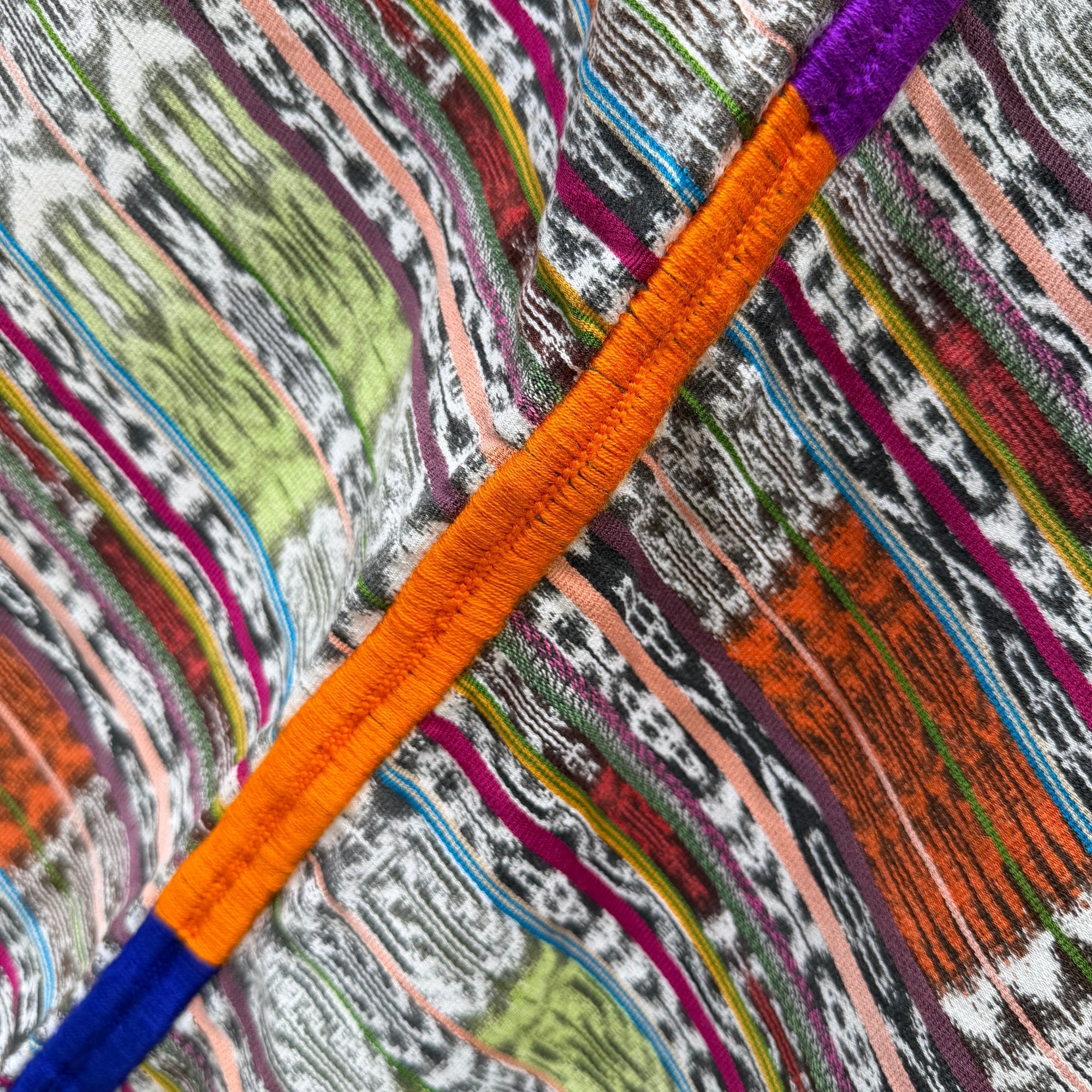 Close-up of a colorful patterned textile with abstract wavy stripes in orange, purple, blue, green, pink, black, and white, overlaid with a prominent orange embroidered line running diagonally across the frame.