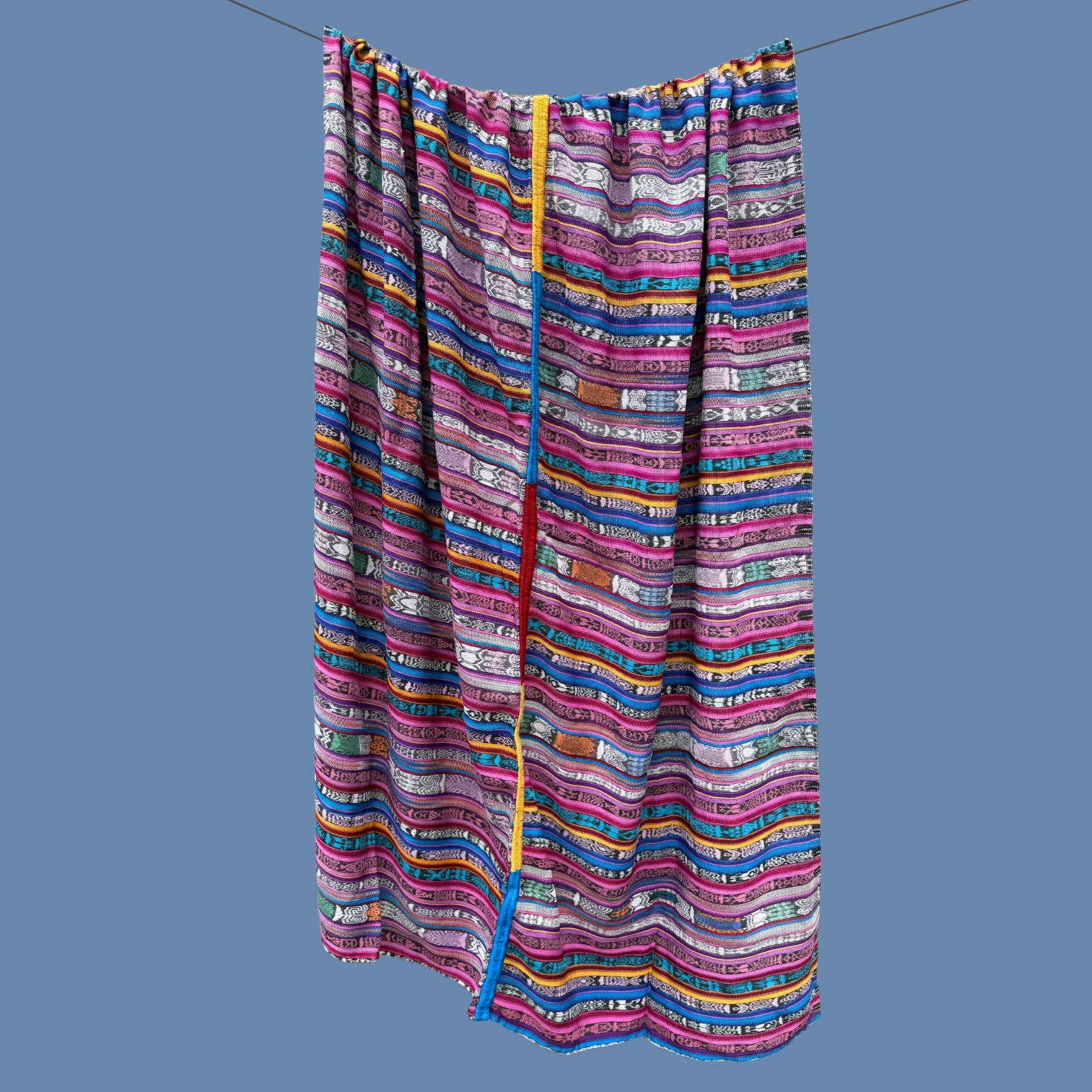 A colorful striped textile hanging from a wire against a solid blue-gray background. The fabric features horizontal bands of pink, purple, blue, yellow, orange, and white with a central vertical embroidered stripe in red and yellow.