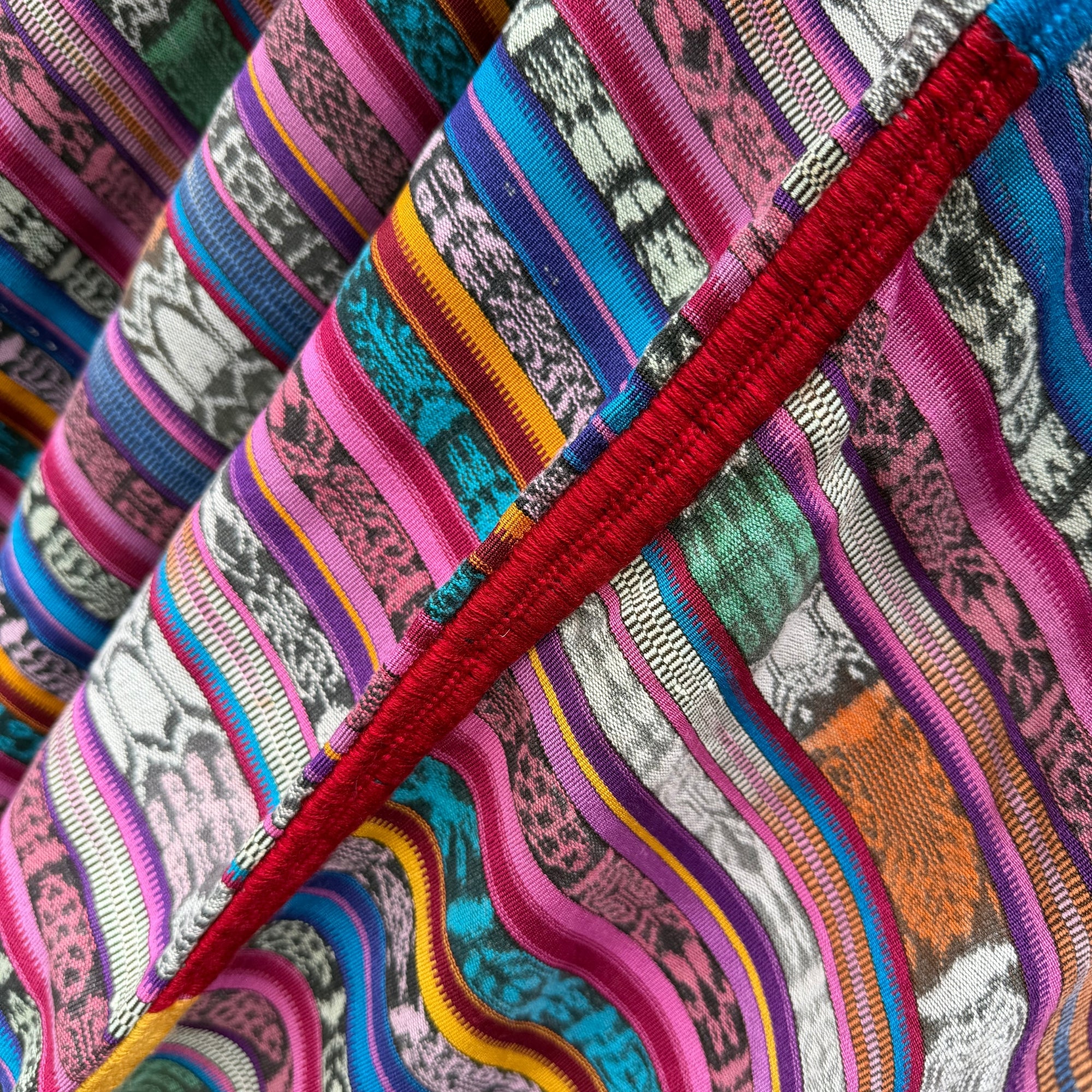 Close-up of a colorful striped textile with woven patterns in pink, blue, teal, orange, yellow, black, and white, crossed by a prominent red embroidered seam.