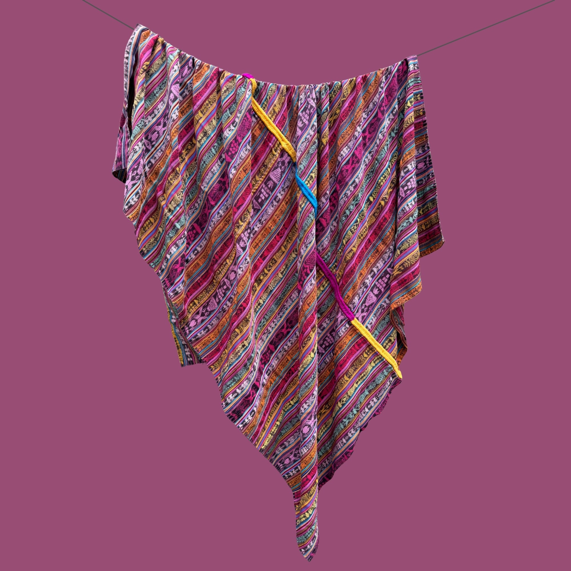 A multicolor patterned blanket with diagonal stripes in pink, purple, orange, yellow, and green, hanging from a thin wire against a solid mauve background.