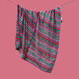 A patterned blanket with diagonal stripes in green, pink, red, and white, hanging from a black clothesline against a solid pink background.