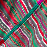 Close-up of a colorful striped textile with wavy vertical bands in green, red, pink, gray, white, and black, crossed by a prominent green stitched seam.