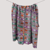 A multicolor patterned throw blanket with a ikat design in purple, green, orange, and white, hanging from a clothesline against a solid light gray background.