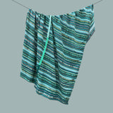 A throw blanket with an ikat horizontal striped pattern in shades of teal, green, blue, and beige, hanging from a thin wire against a solid gray background.