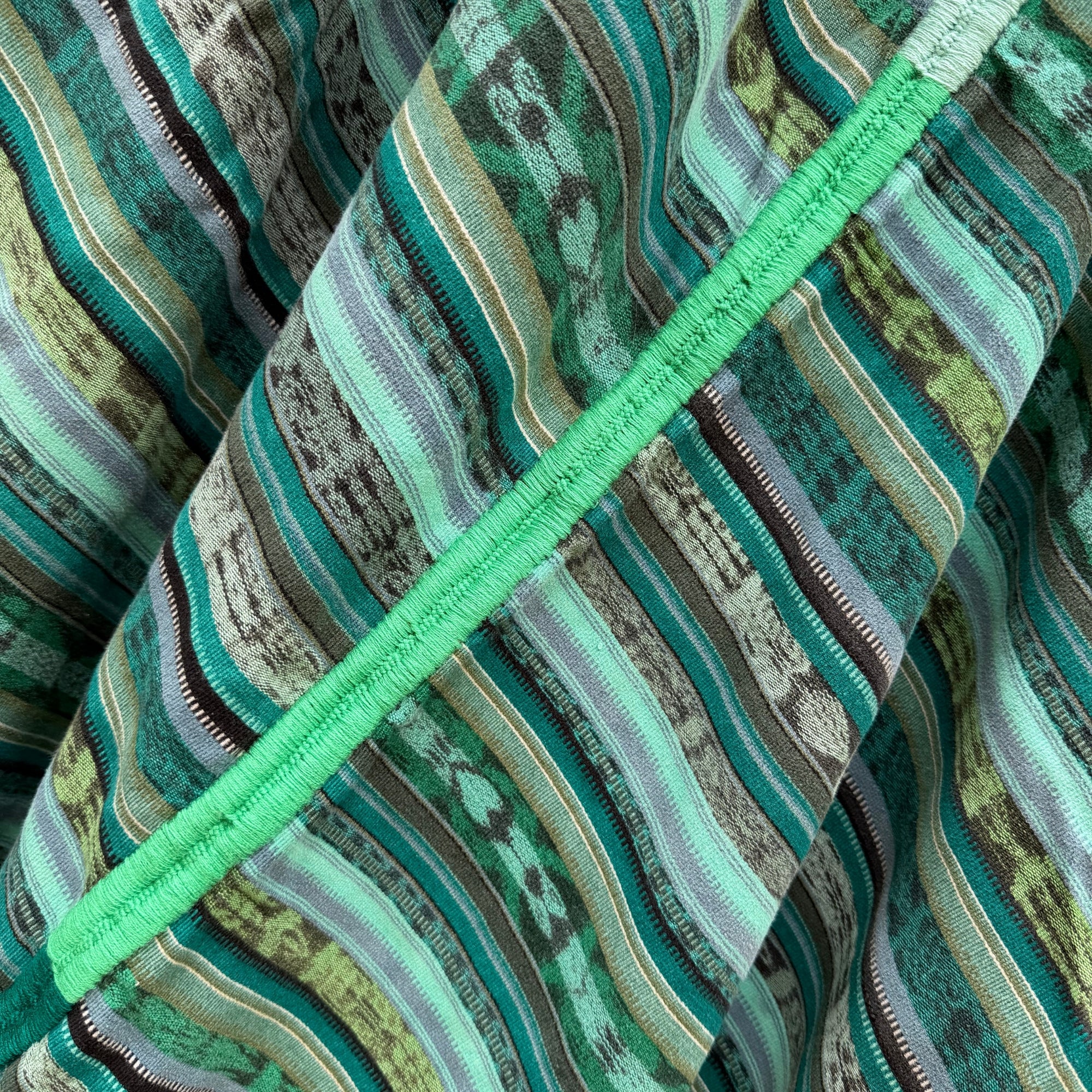 Close-up of a striped woven textile in shades of green, teal, gray, and beige, with a bright mint green stitched trim running diagonally across the frame.