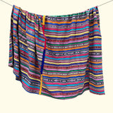 A colorful striped fabric throw blanket, hanging from a string against a plain cream background. The fabric features horizontal bands of pink, blue, yellow, green, orange, and black with geometric patterns.