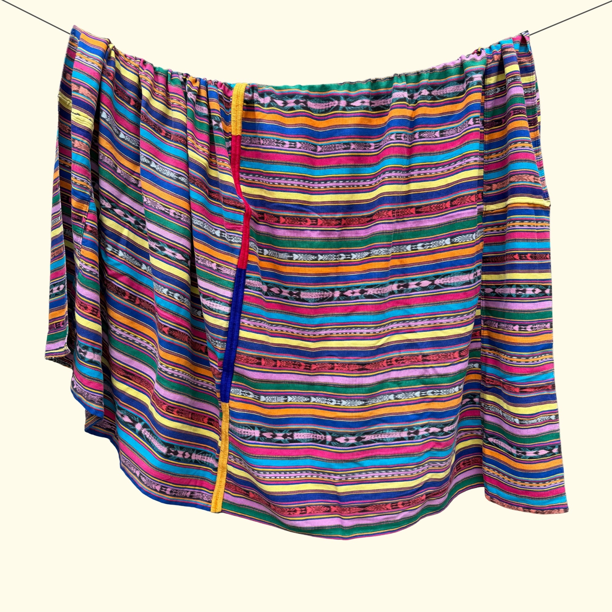 A colorful striped fabric throw blanket, hanging from a string against a plain cream background. The fabric features horizontal bands of pink, blue, yellow, green, orange, and black with geometric patterns.
