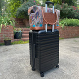 A multicolored geometric-patterned overnight travel bag with a brown leather base and a black trolley sleeve, placed on the ground next to a suitcase.