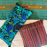 Two decorative throw pillows displayed on a bed with a colorful woven textile behind them. One pillow features a bold blue and green floral pattern on a black background, and the other features a vibrant multicolor striped woven design.