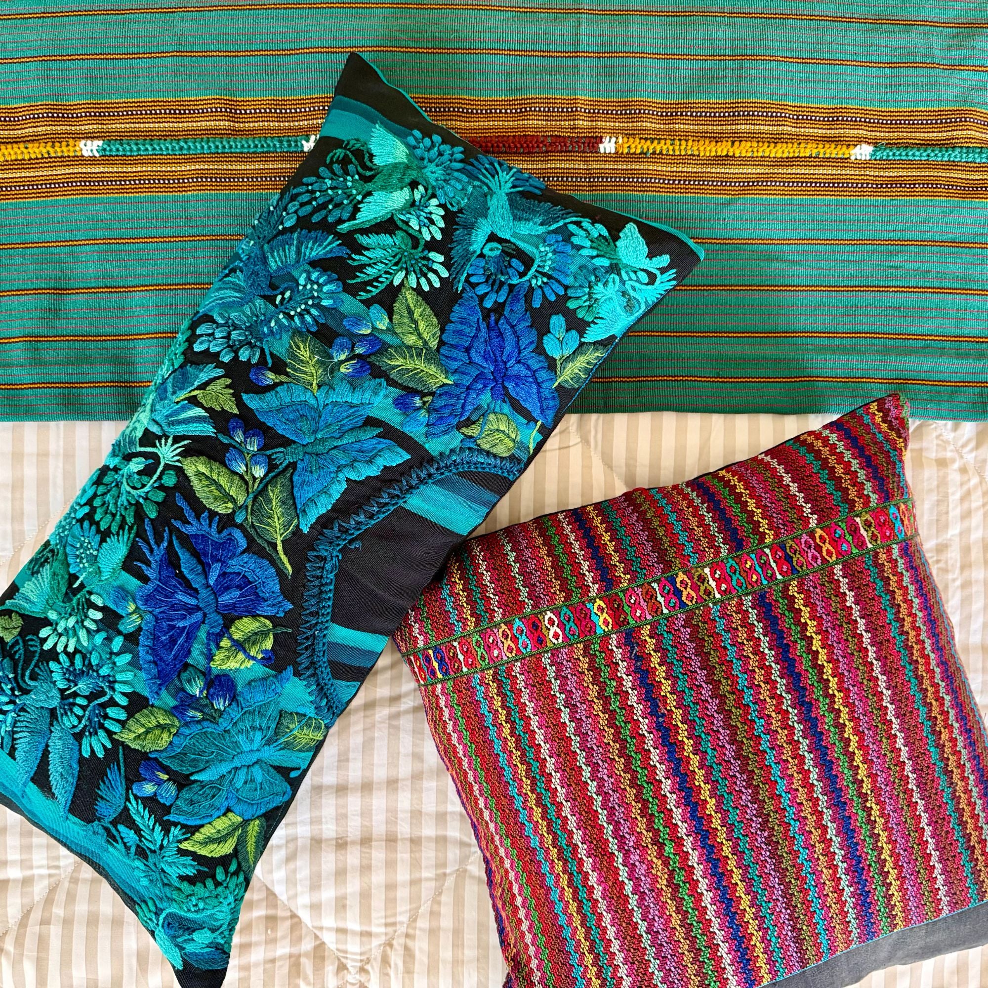 Two decorative throw pillows displayed on a bed with a colorful woven textile behind them. One pillow features a bold blue and green floral pattern on a black background, and the other features a vibrant multicolor striped woven design.