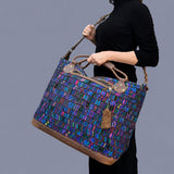 A person in a black outfit carries a large tote bag with a vibrant, multicolored geometric pattern and brown leather handles against a solid gray background.