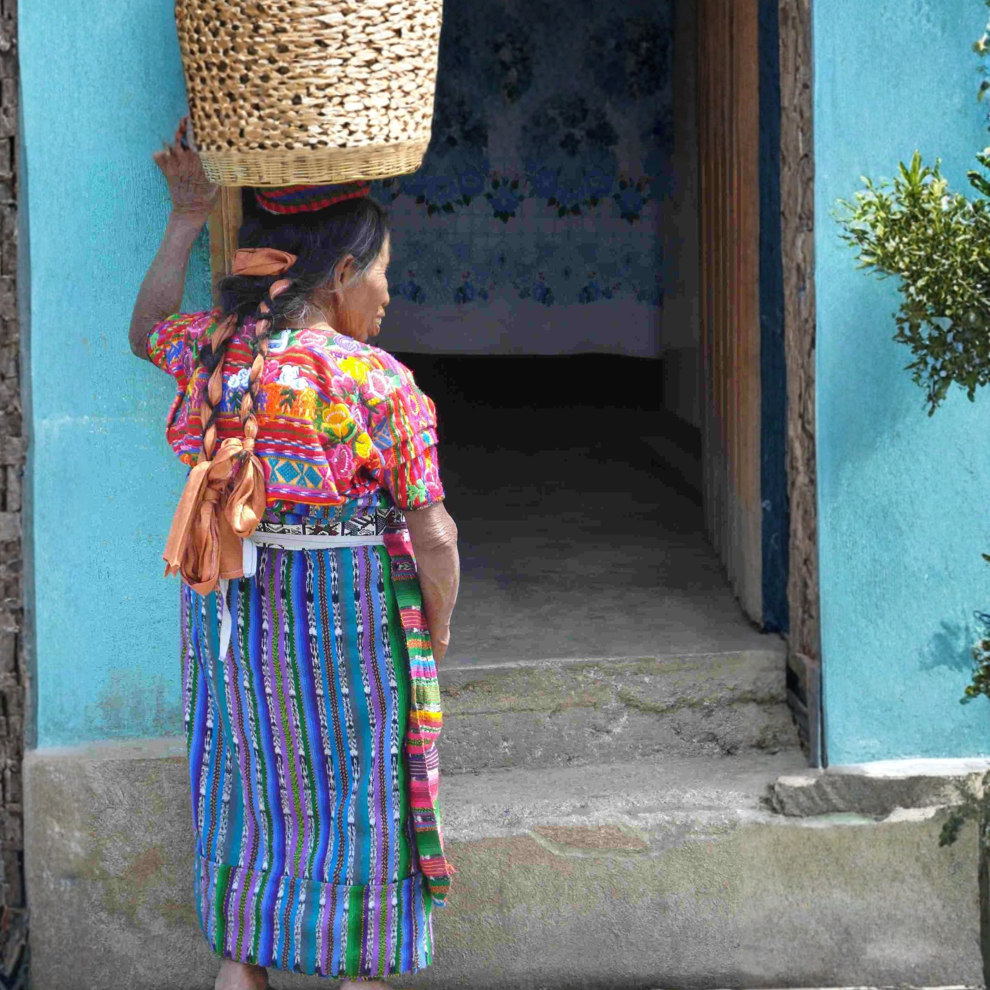 Woman in traditional colorful dress carrying a woven basket, standing in front of a blue wall.