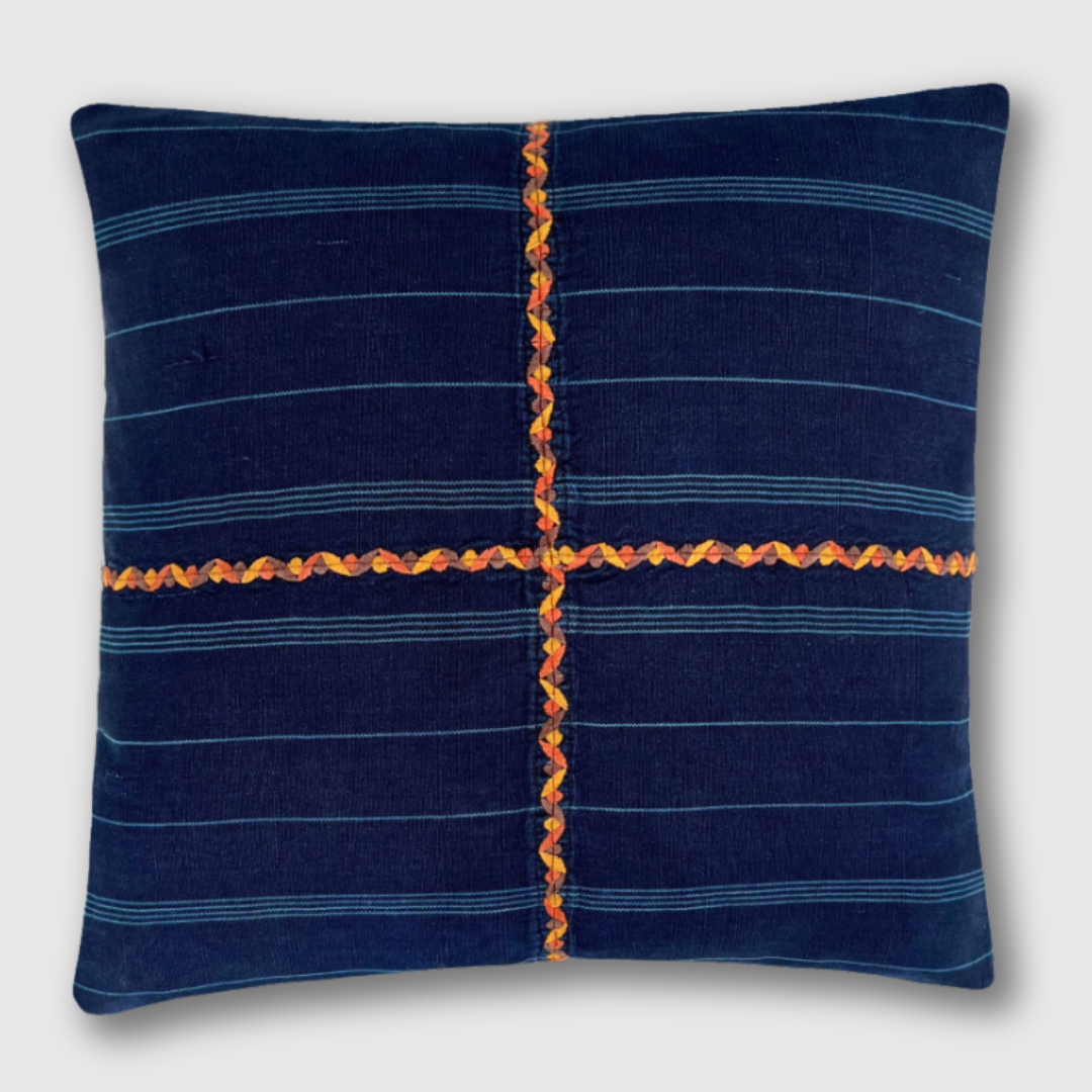 Indigo corte pillow on a light gray background with an orange embroidered cross on the front. 