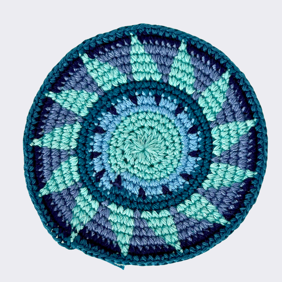 Close up of blue, green, and black crocheted coaster on light gray background - Intertwined: Handmade for Good
