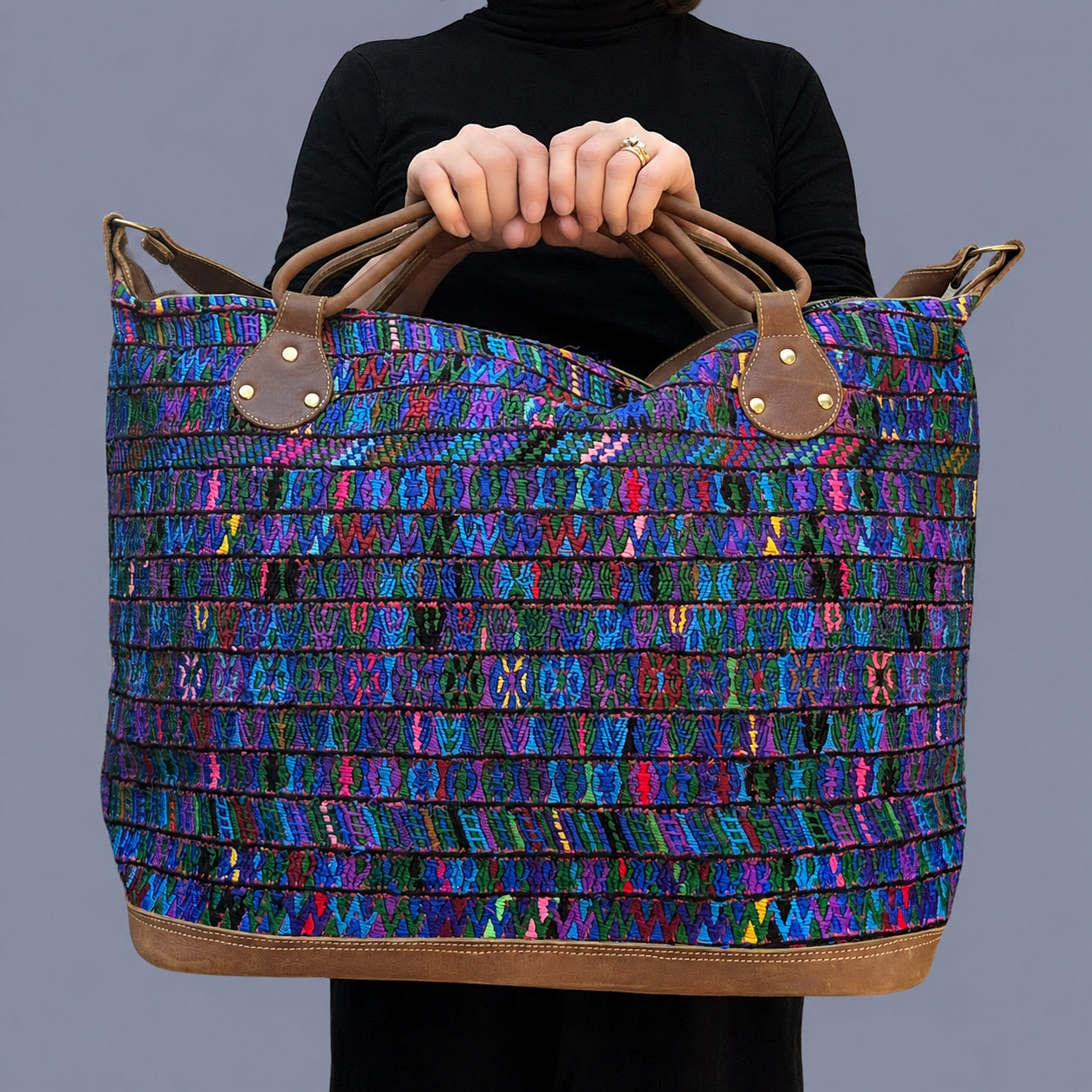 A person holding a large woven tote bag with a vibrant multicolor pattern of blue, purple, pink, and green threads, featuring brown leather handles and trim against a solid gray background.