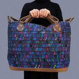 A person holding a large woven tote bag with a vibrant multicolor pattern of blue, purple, pink, and green threads, featuring brown leather handles and trim against a solid gray background.
